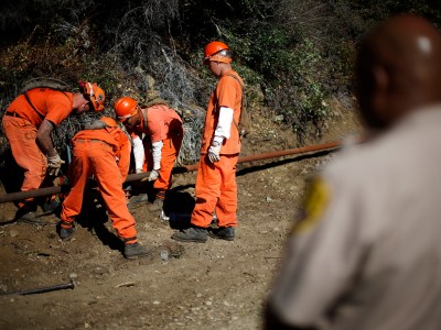 A group of inmates dressed in orange uniforms and helmets are working together outdoors, bending down to manipulate a long metal rod in a dirt area surrounded by trees. In the foreground, a person in a beige police uniform is slightly blurred, observing the scene.